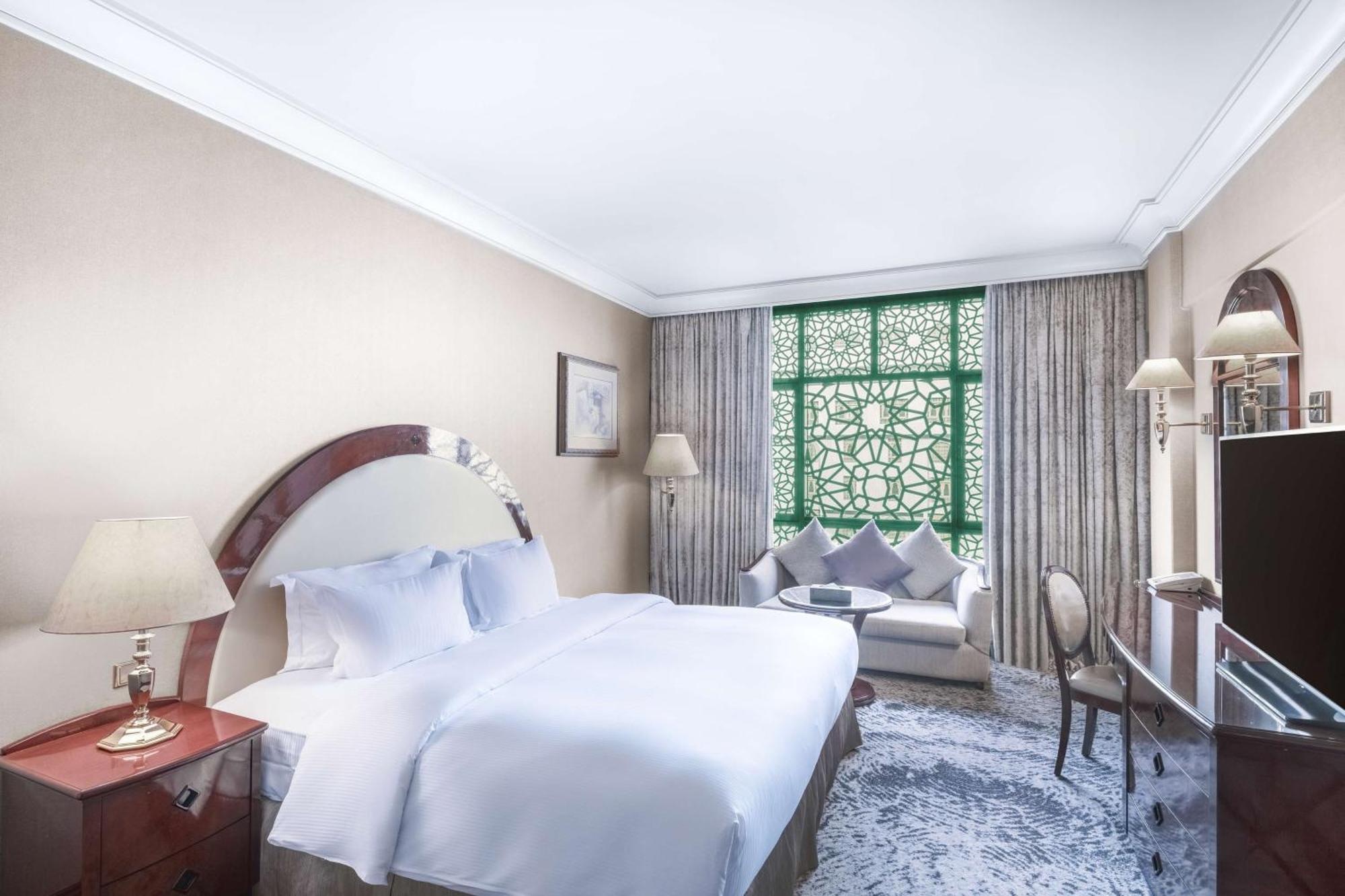 Madinah Hilton Hotel Medina Exterior photo The photo depicts a luxurious hotel room featuring a large bed with white bedding, flanked by bedside lamps. There are elegant curtains lining the window that includes a decorative green lattice design. In the corner of the room, there is a cozy seat