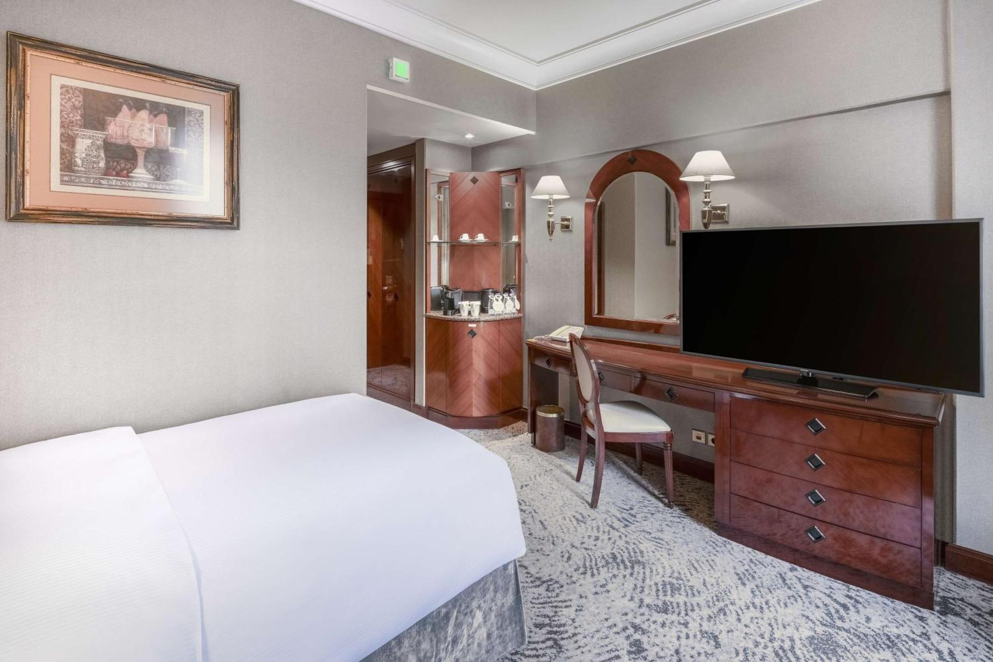 Madinah Hilton Hotel Medina Exterior photo The photo shows an elegantly designed hotel room. It features a neatly made bed with a white duvet, a wooden desk with a chair, and a large flat-screen television. There is a dresser with drawers and decorative elements, including a mirror. The walls