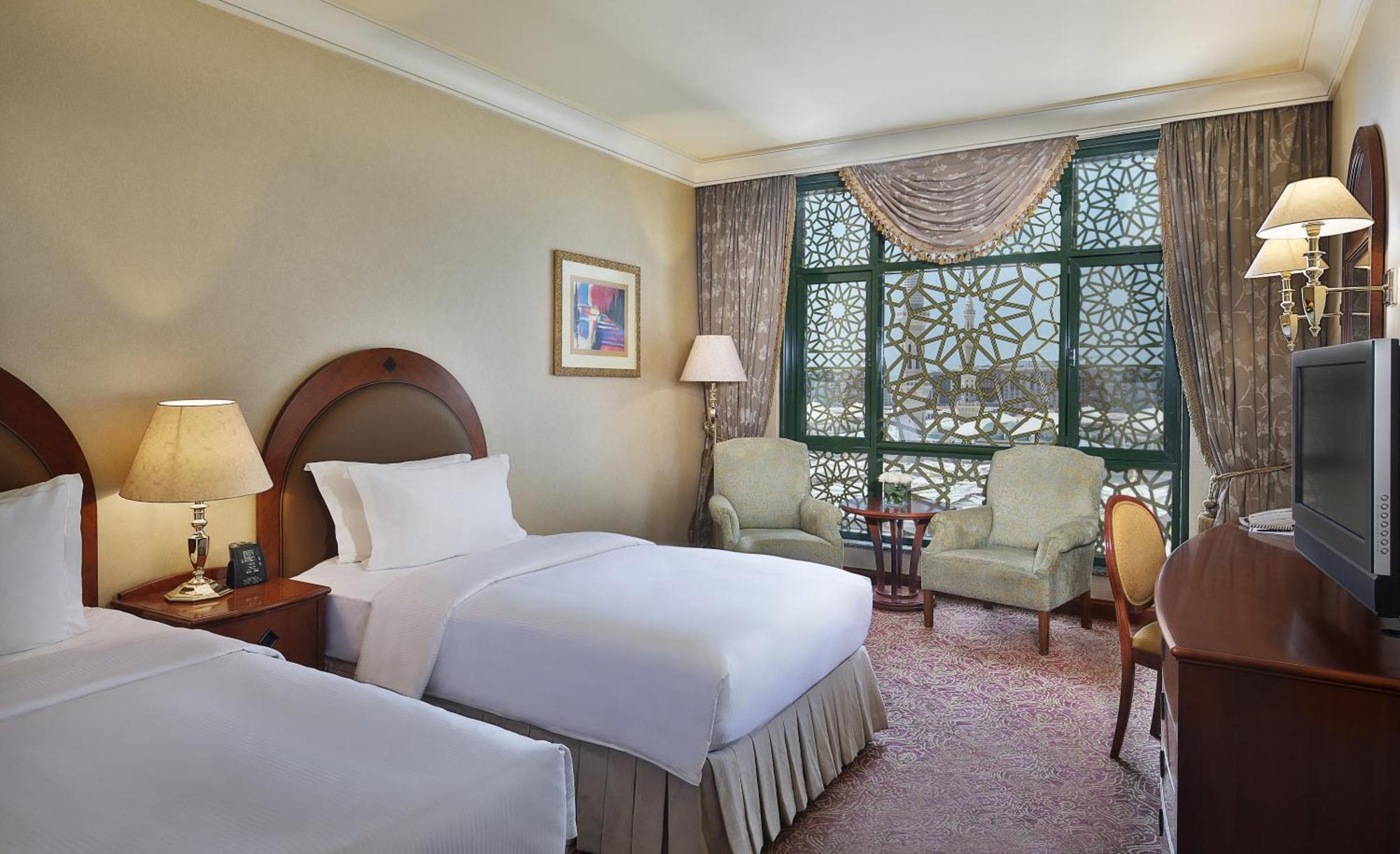 Madinah Hilton Hotel Medina Exterior photo The photo shows a cozy hotel room that features two twin beds with white linens positioned against a beige wall. There's a wooden bedside table with a lamp next to one of the beds. Across the room, there are two comfortable chairs and a small round t