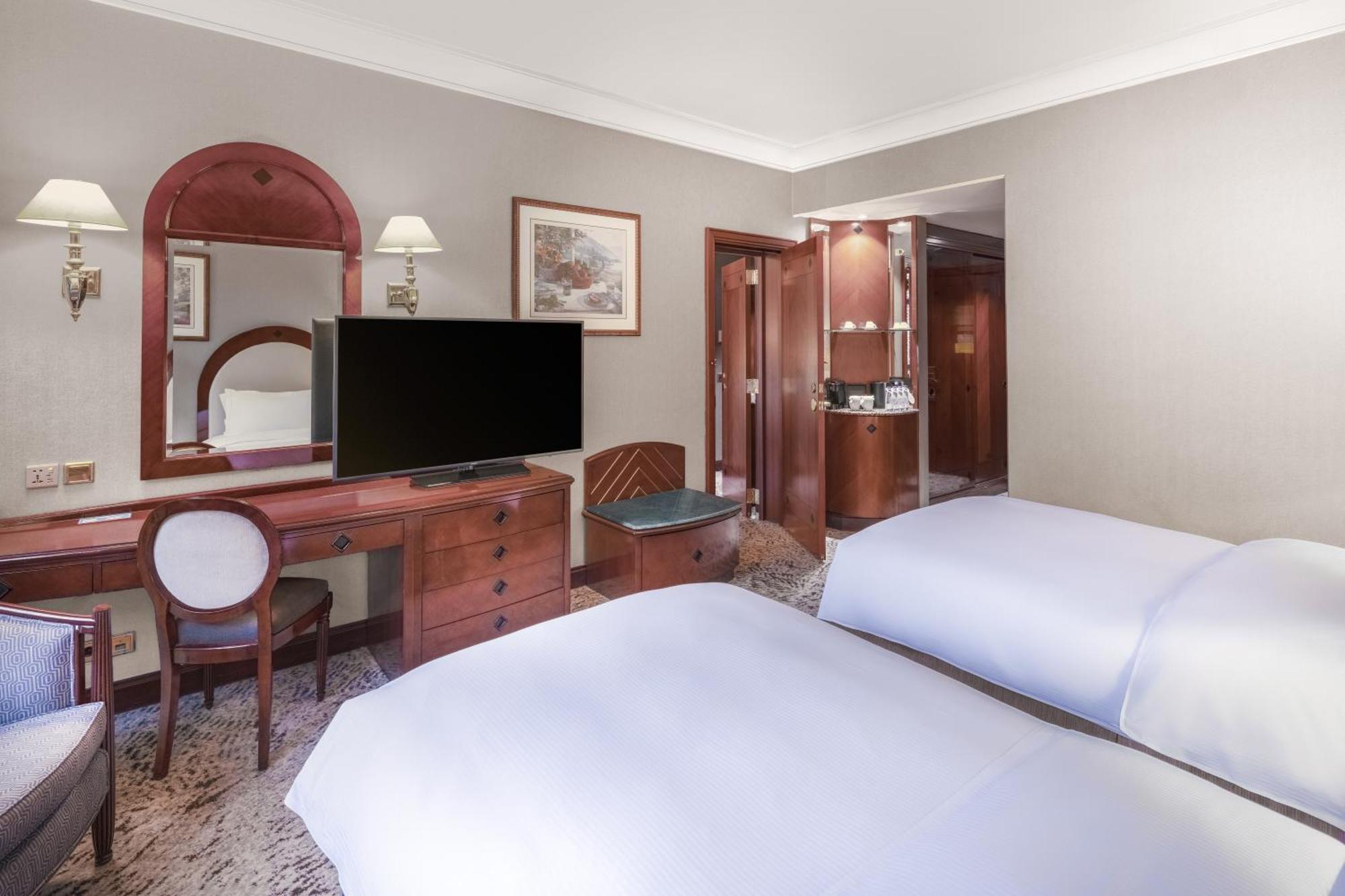 Madinah Hilton Hotel Medina Exterior photo The photo shows an interior of a hotel room. The room features two beds with white bedding. There's a wooden desk with a chair and a large TV mounted on the wall. A mirror is placed above the desk, and there are bedside lamps on either side of the be