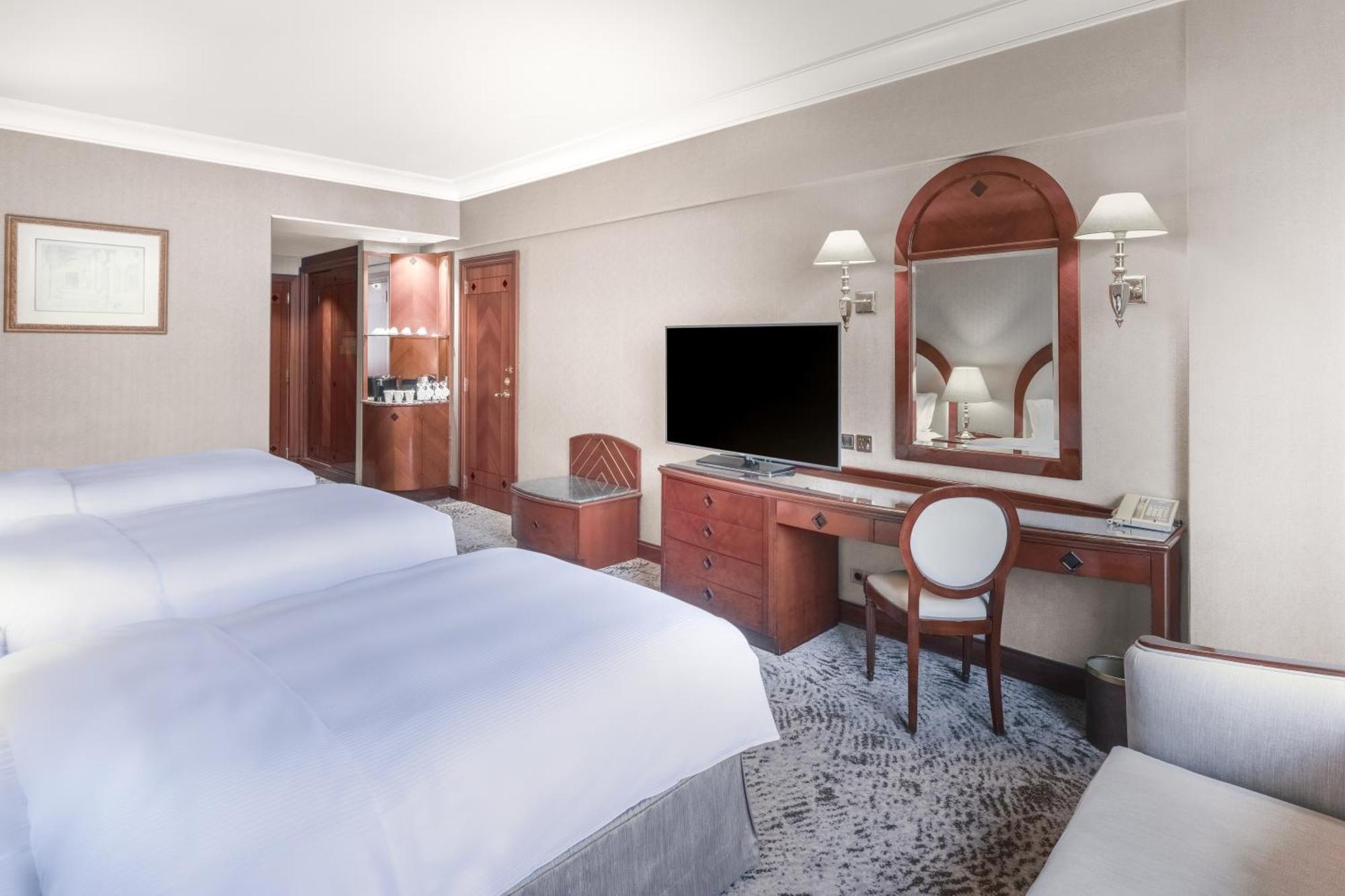 Madinah Hilton Hotel Medina Exterior photo The photo shows a hotel room with two twin beds, both made with white bedding. There is a wooden dresser with a TV mounted on the wall above it. A mirror is positioned above the dresser, and there is a small armchair and a decorative rug on the floor