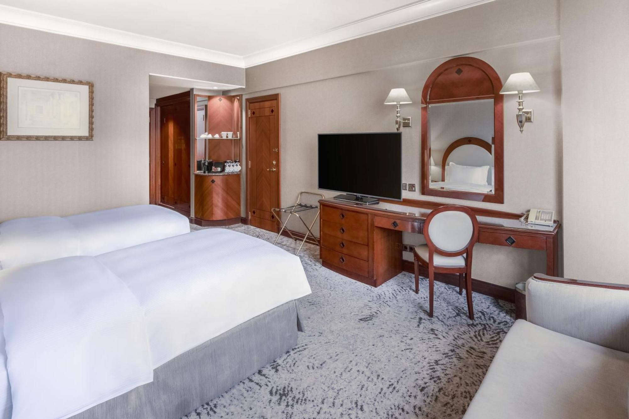 Madinah Hilton Hotel Medina Exterior photo The photo shows a well-designed hotel room featuring two single beds with white bedding. There is a wall-mounted flat-screen TV across from the beds. A wooden desk and chair are situated next to the TV, with a mirror hanging above the desk. Near the 
