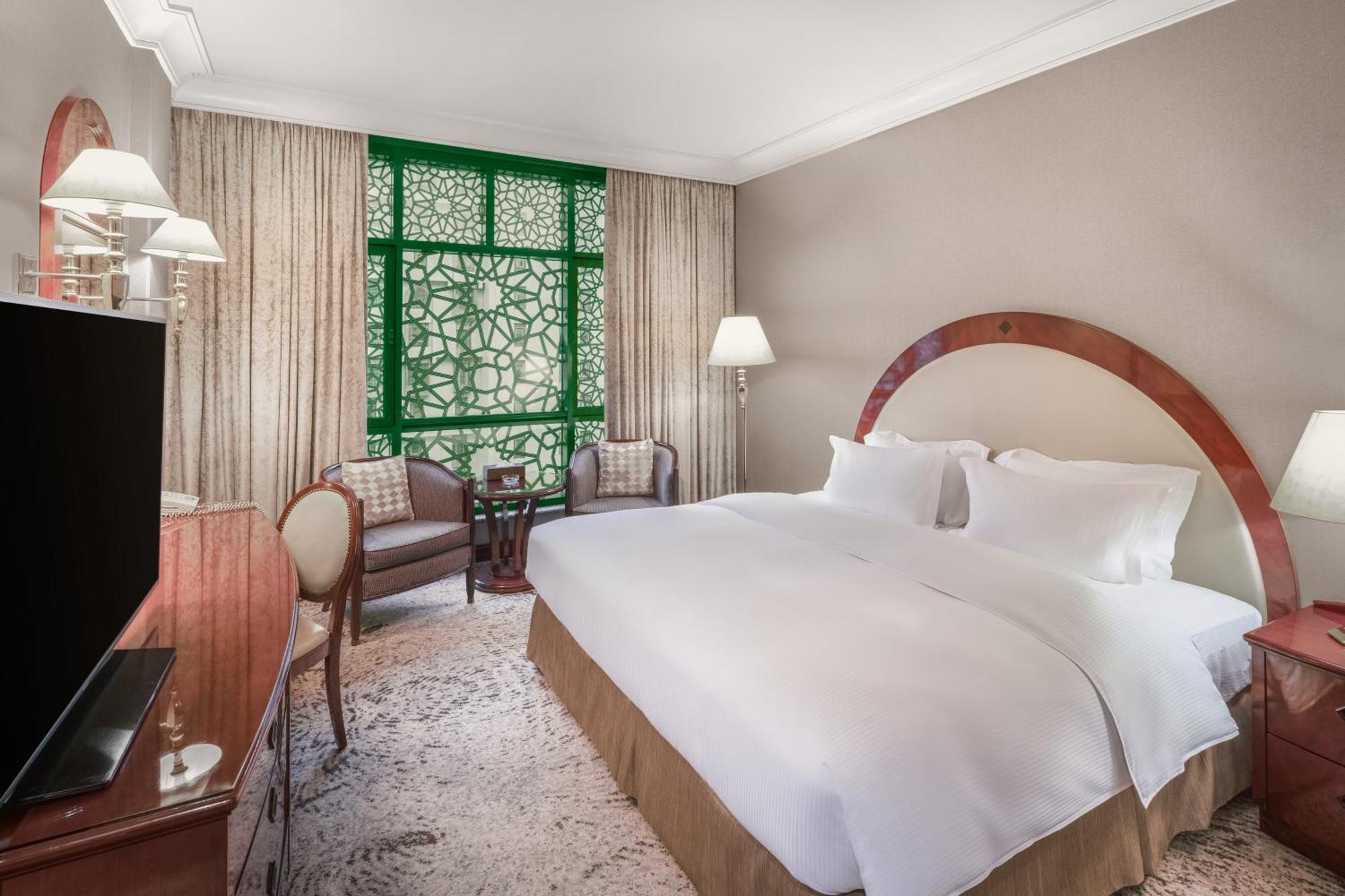 Madinah Hilton Hotel Medina Exterior photo The photo shows a beautifully designed hotel room featuring a large king-sized bed with white linens. The room is decorated in neutral tones, giving it a warm and inviting atmosphere. 

To one side, there's a wooden dresser with a television on top. 