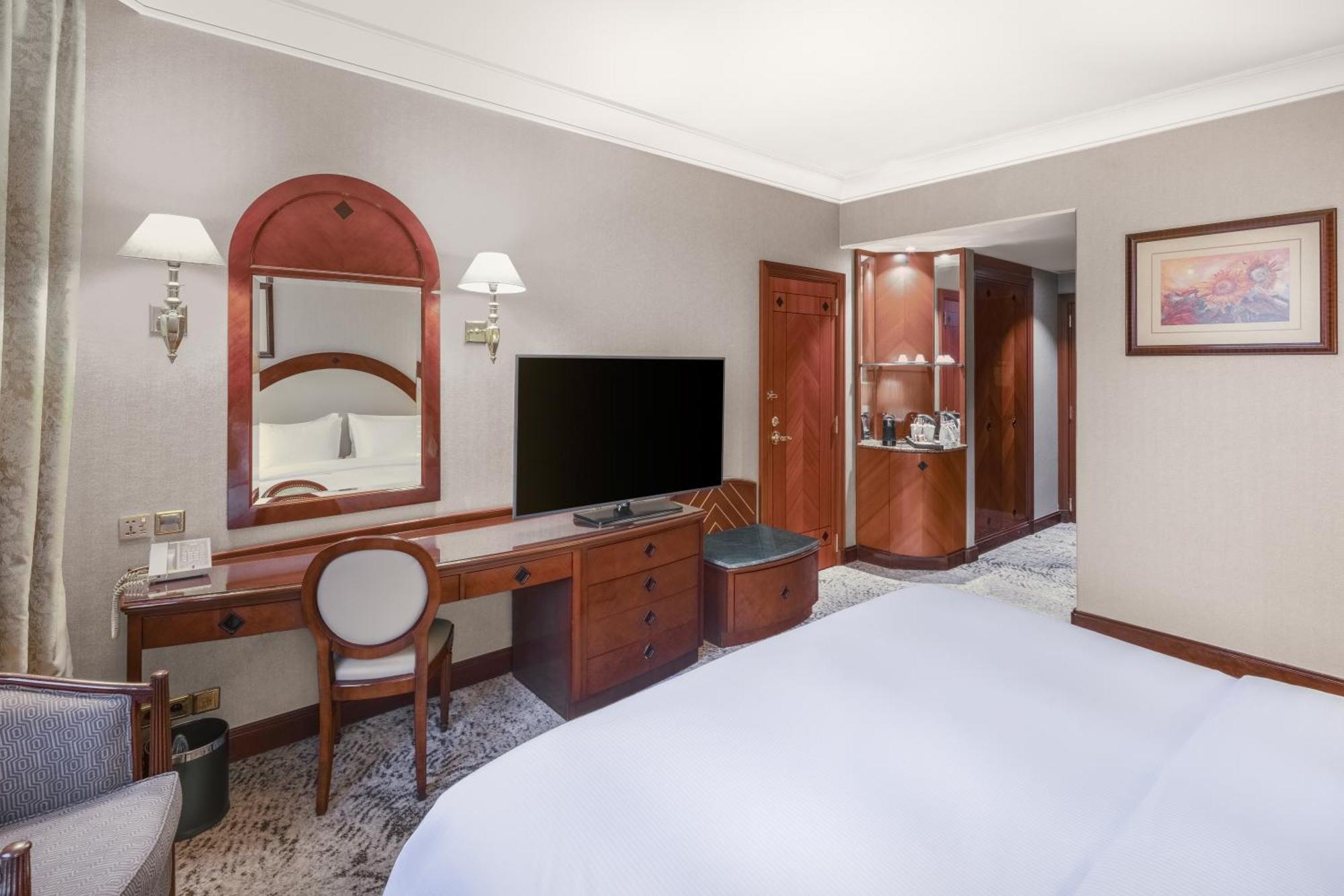 Madinah Hilton Hotel Medina Exterior photo The photo shows a hotel room that features a neatly made bed with white linens. To one side, there is a wooden desk with a round chair and a large mirror above it. A flat-screen TV is mounted on the wall opposite the bed. The room has soft lighting f