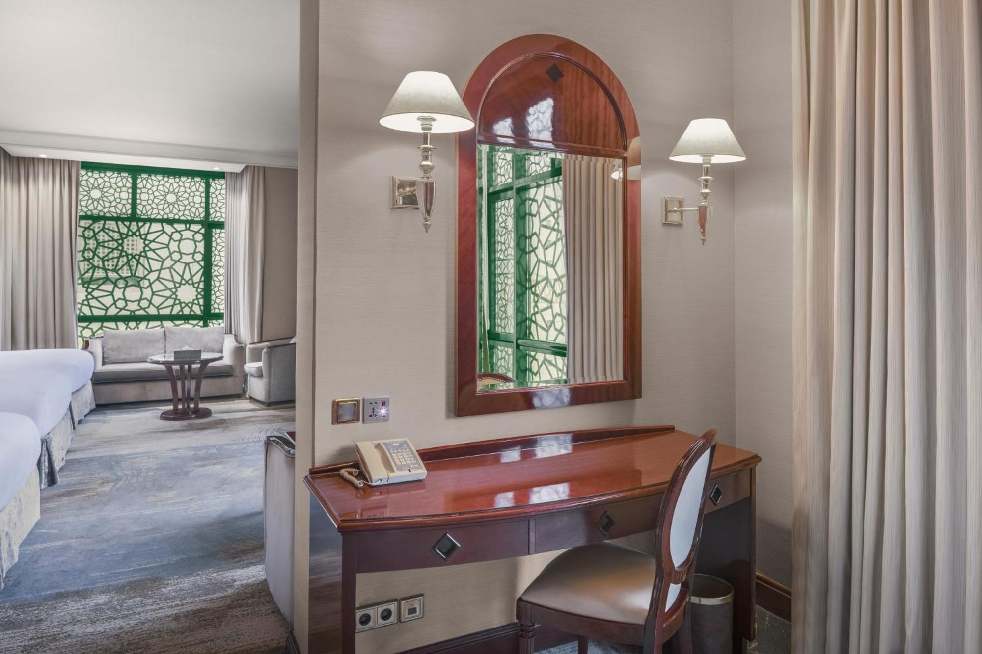 Madinah Hilton Hotel Medina Exterior photo The photo shows a well-furnished hotel room or suite. On the left, there is a wooden vanity desk with a mirror above it, accompanied by a chair. The desk features a phone. Behind the desk, there are curved arch windows with intricate green designs th