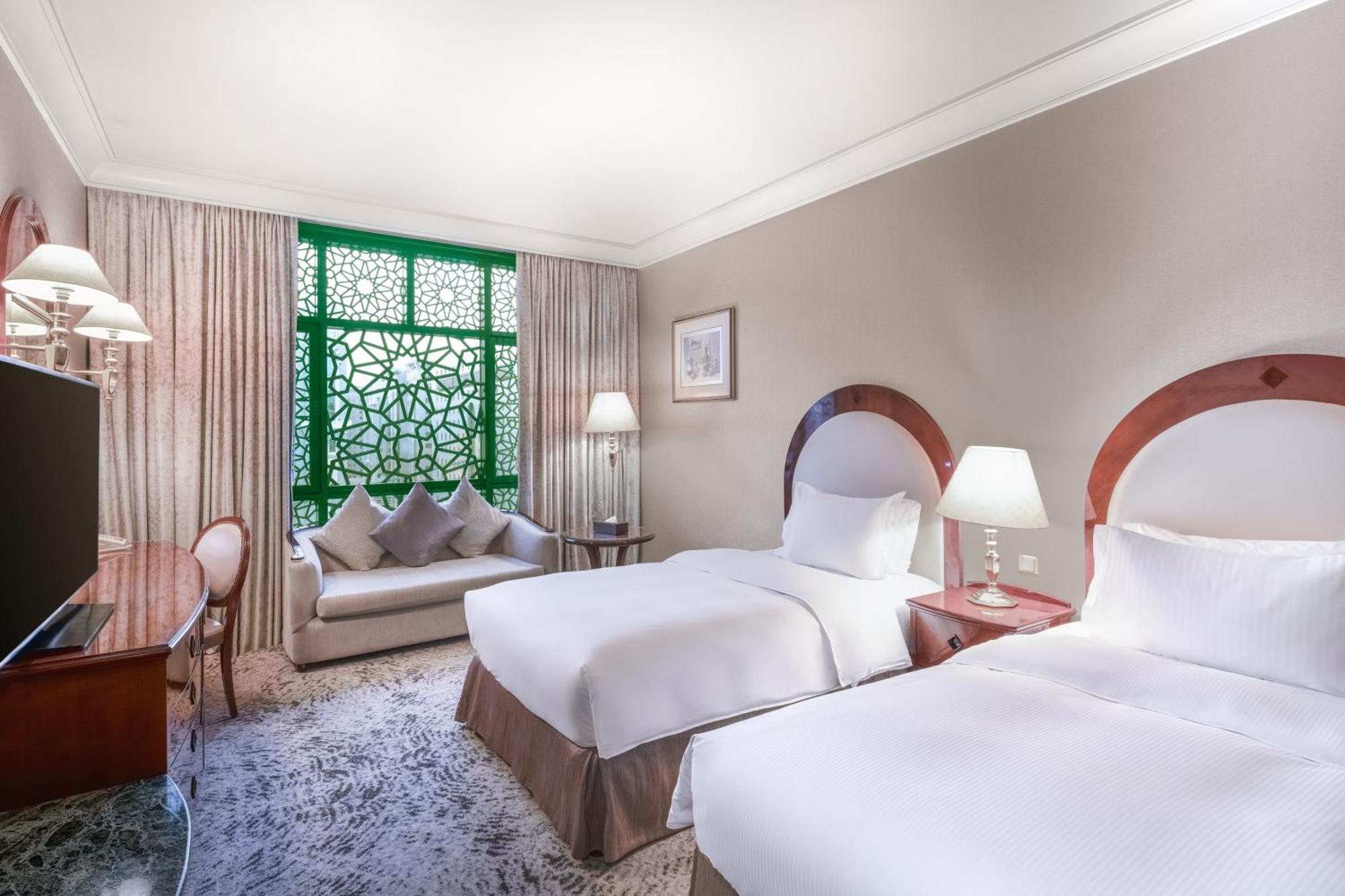 Madinah Hilton Hotel Medina Exterior photo The photo shows a hotel room featuring two twin beds with white linens, a comfortable seating area with a sofa, and a desk with a chair. The room is well-lit with lamps placed beside the beds and on the desk. There is a large window with decorative g