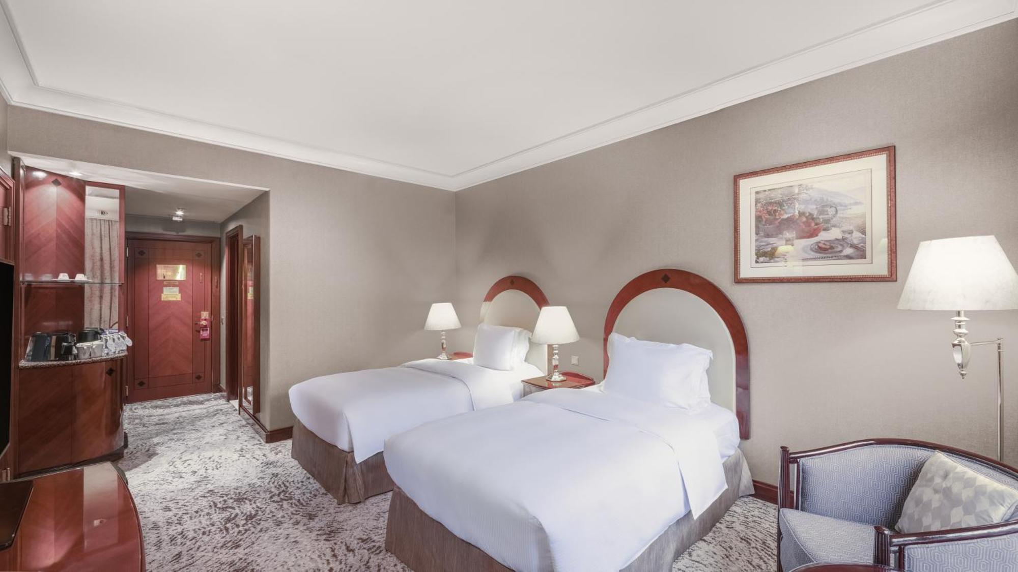 Madinah Hilton Hotel Medina Exterior photo The photo shows a hotel room with a warm and inviting decor. It features two twin beds with white linens and light-colored bedspreads. Each bed has a small bedside table with a lamp next to it. The walls are painted in a neutral tone, and there is a 