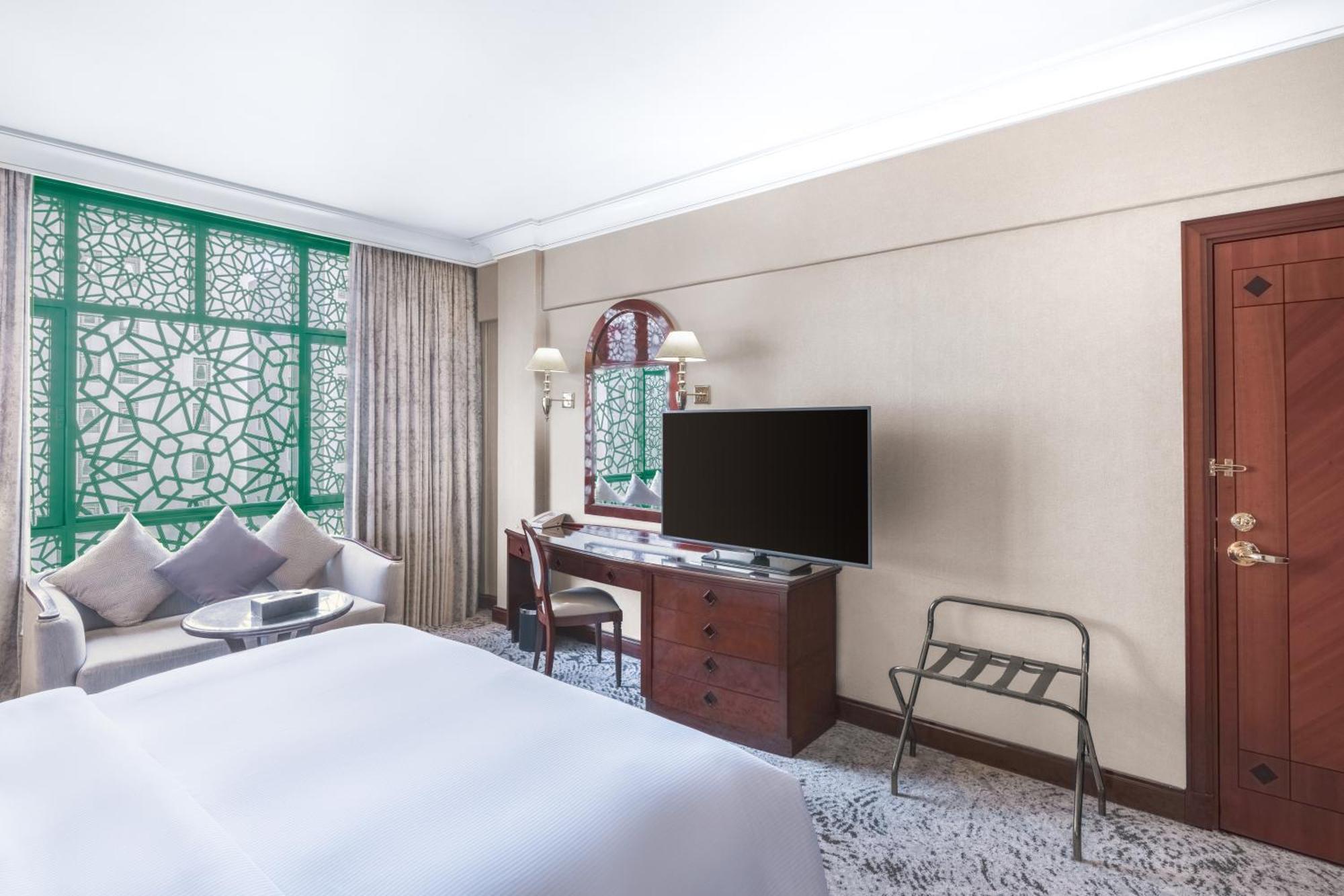 Madinah Hilton Hotel Medina Exterior photo The photo shows a hotel room with a spacious and elegant design. It features a large bed covered with white linens. To one side, there are decorative cushions on a small seating area that includes a round table. A modern television is mounted on a wo
