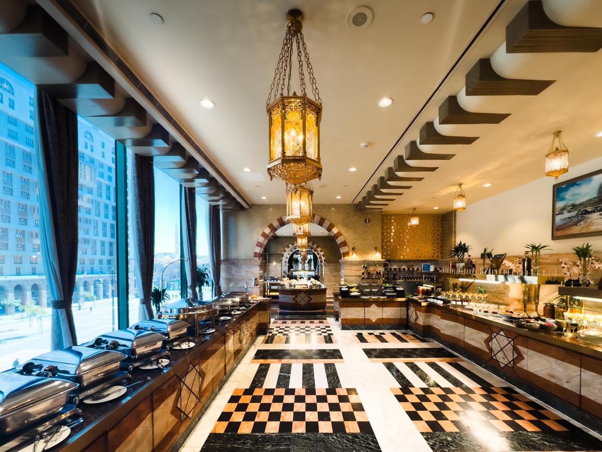 Madinah Hilton Hotel Medina Exterior photo The image shows an elegant buffet restaurant interior. The space features a stylish chandelier hanging from the ceiling, providing warm lighting. There are large windows on one side, allowing natural light to enter. The floor has a distinctive black 