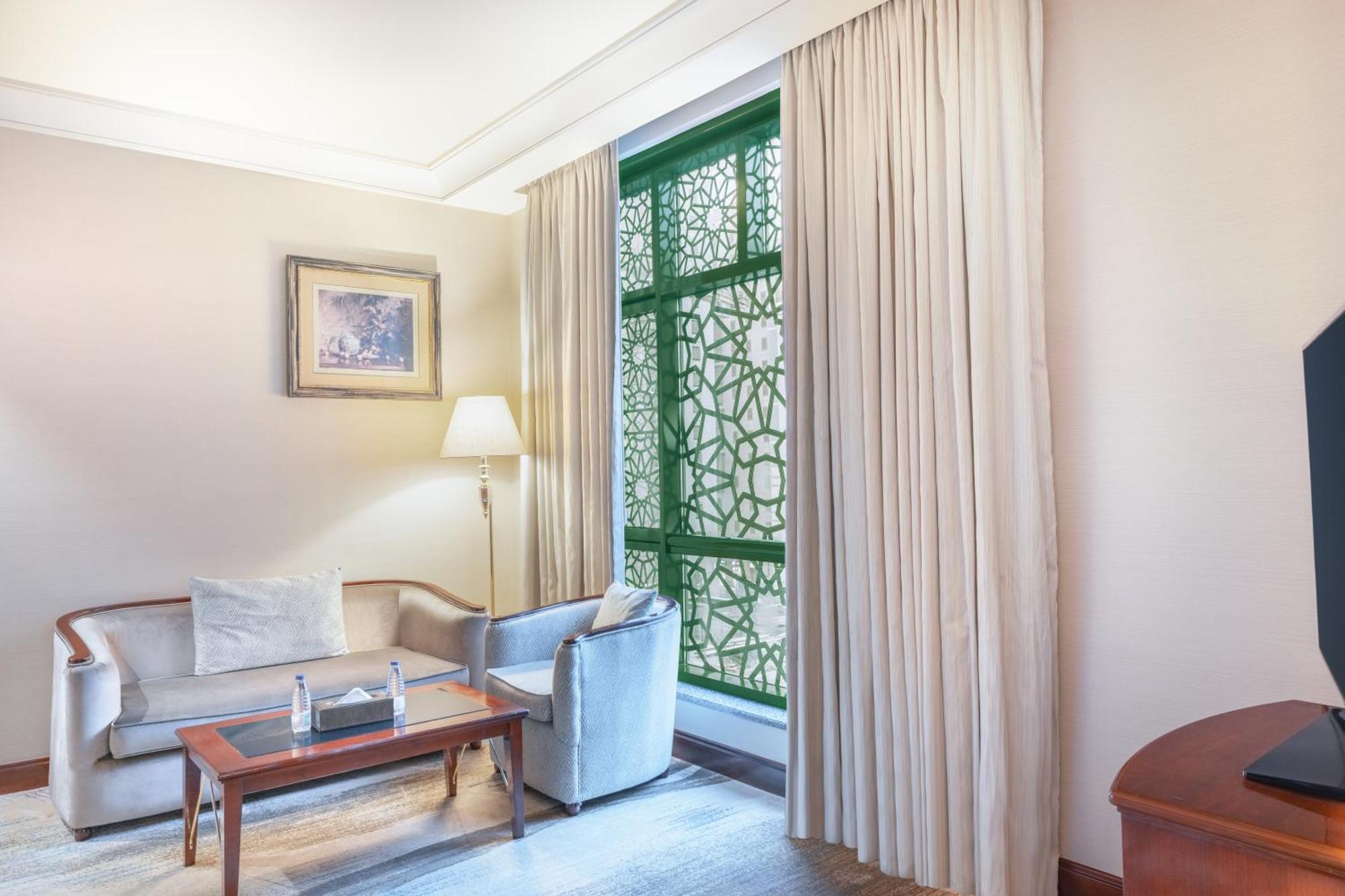 Madinah Hilton Hotel Medina Exterior photo The photo shows an interior space, likely a hotel or a private room. It features a light-colored wall and a soft carpet. In the center, there is a small wooden table with two bottles of water. A comfortable couch with cushions is positioned near the 