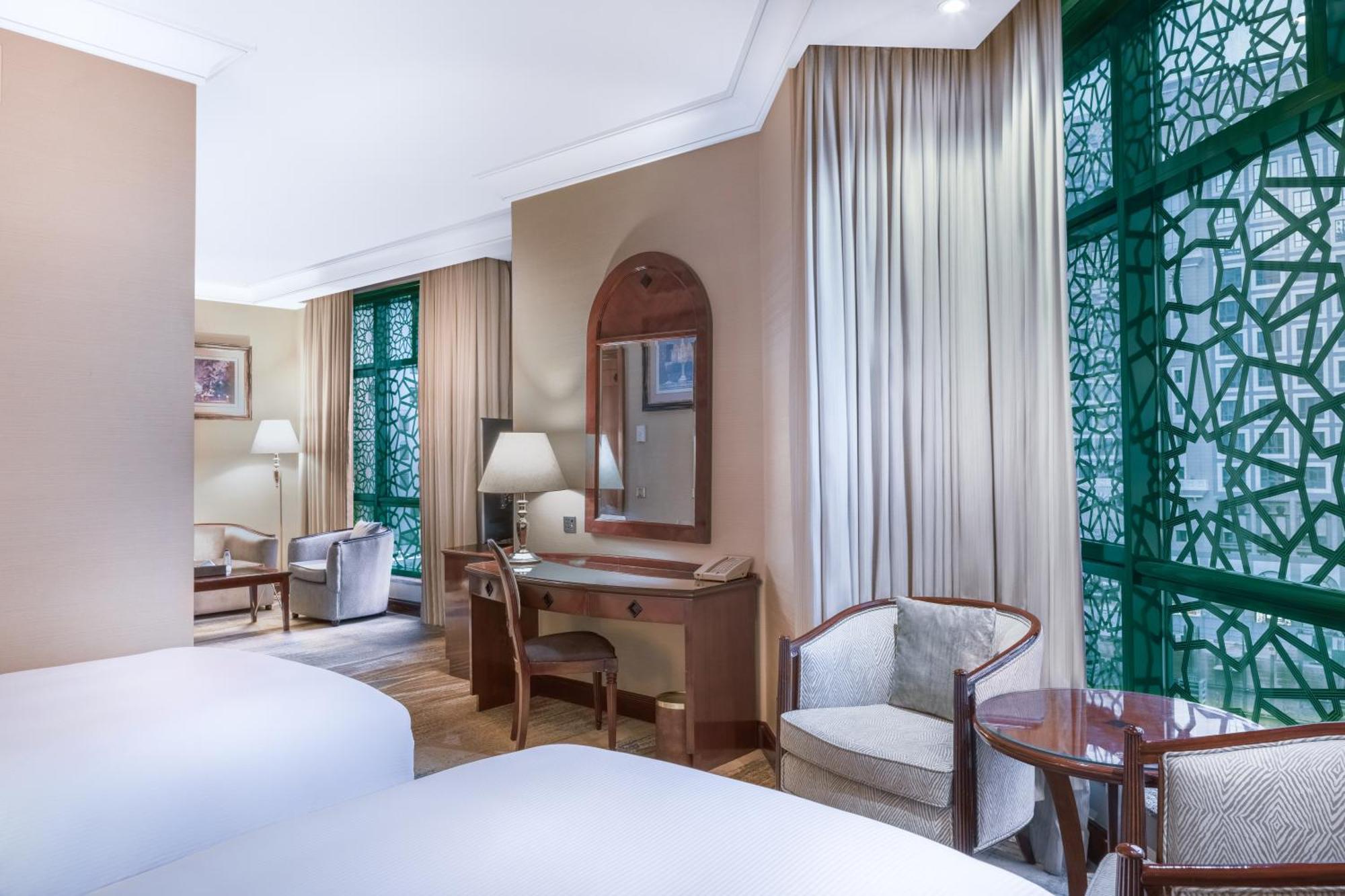 Madinah Hilton Hotel Medina Exterior photo The photo shows a hotel room with a modern and elegant design. It features two beds in the foreground, a sitting area with two chairs and a small round table, and a desk with a mirror. Large windows adorned with decorative green panels allow light to
