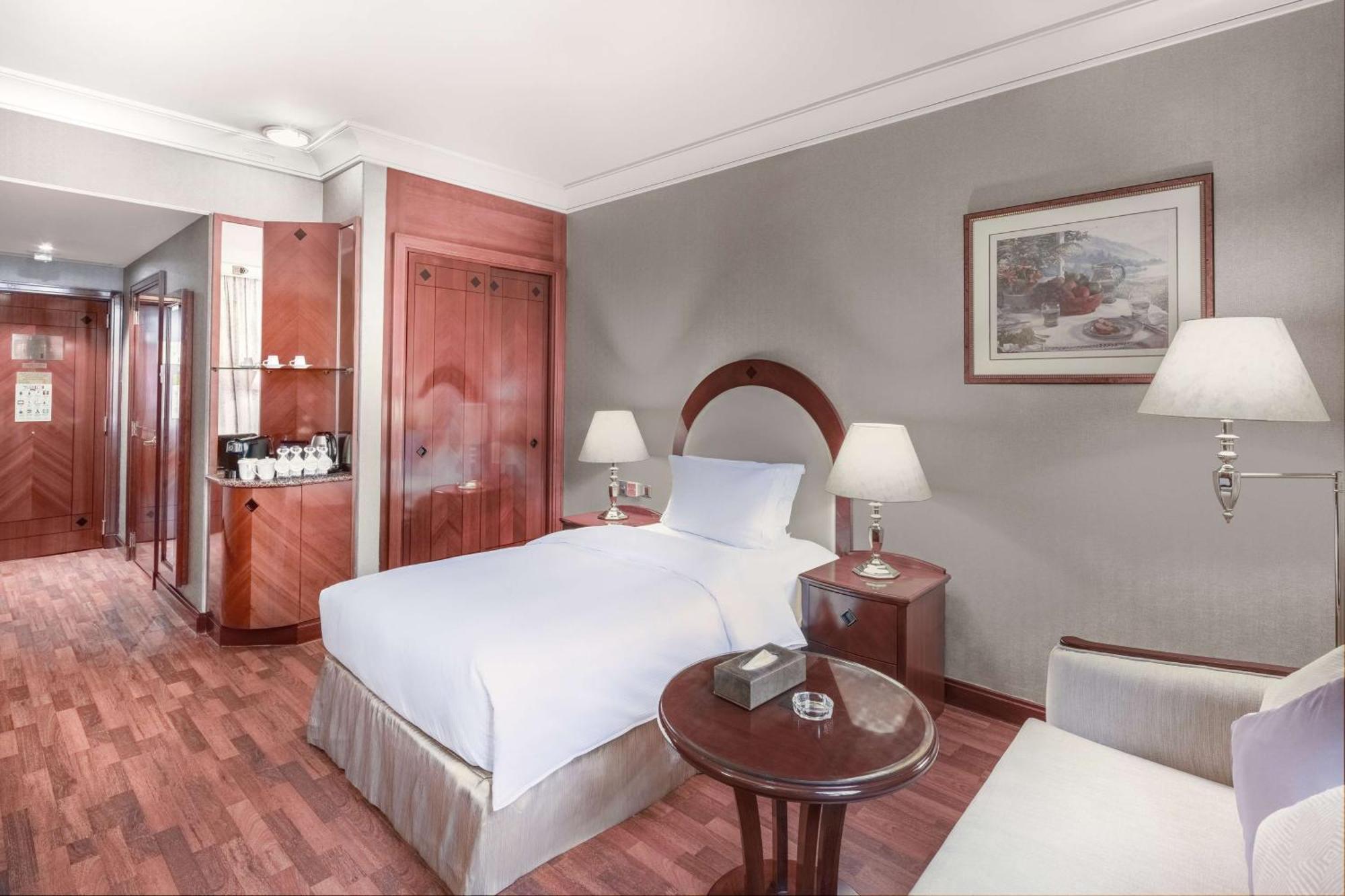Madinah Hilton Hotel Medina Exterior photo The photo shows a well-furnished hotel room with a modern and elegant design. The room features a single bed with white bedding, a small round table next to it, and a comfortable chair. There are bedside lamps on either side of the bed, providing sof