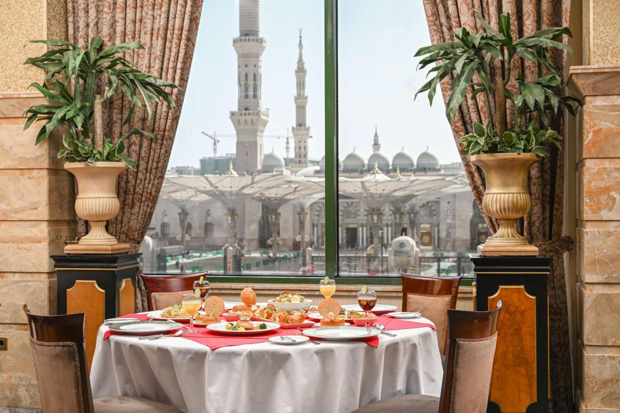 Madinah Hilton Hotel Medina Exterior photo The photo shows an elegantly set dining table in a restaurant, adorned with a tablecloth and plates featuring a variety of dishes. There are glasses and garnishes that suggest a fine dining experience. Large windows provide a view of an impressive ar