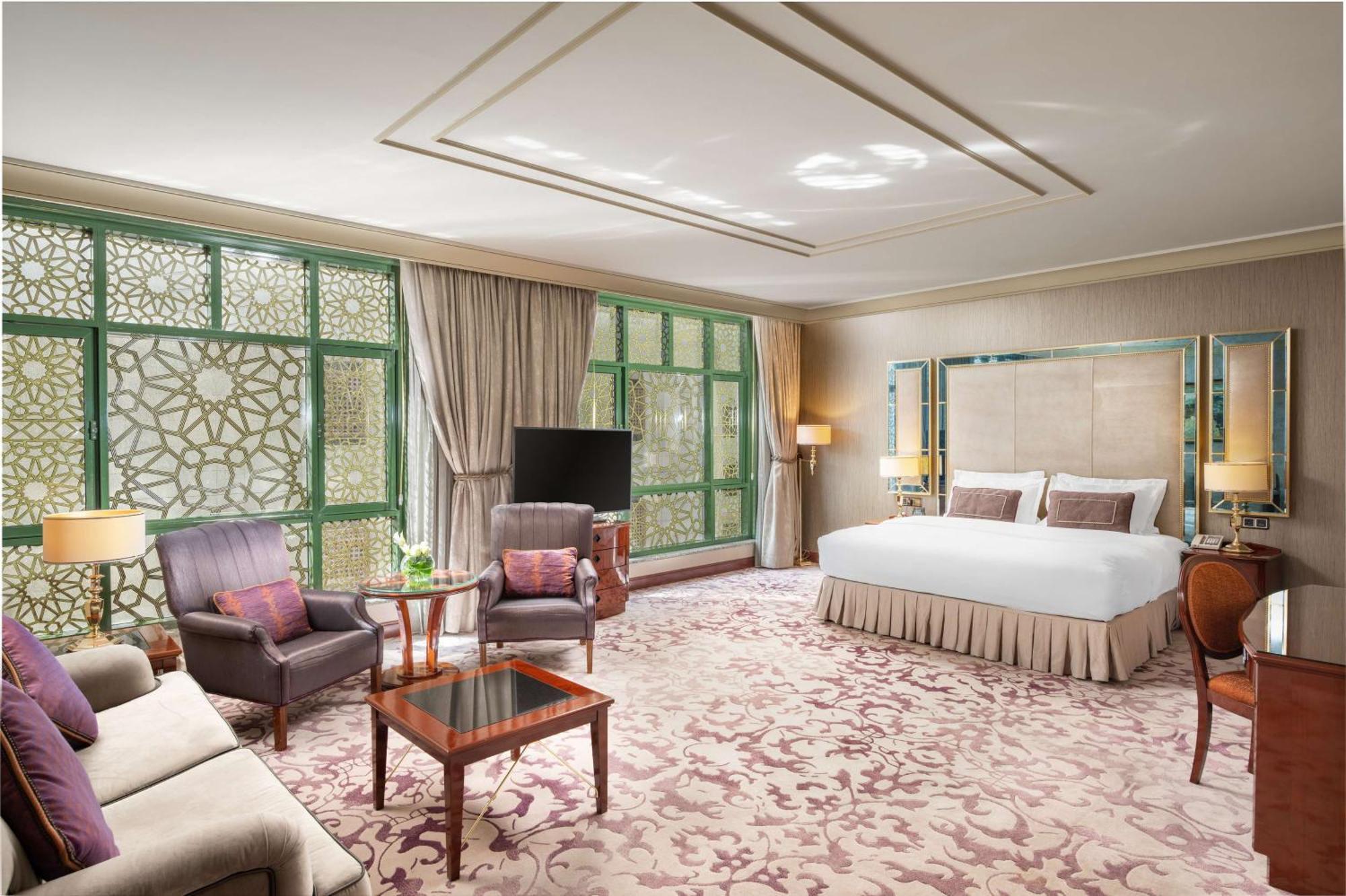 Madinah Hilton Hotel Medina Exterior photo The photo shows a luxurious hotel room featuring a spacious layout. The room includes a large bed with decorative pillows, a cozy seating area with two armchairs and a small table, and a plush sofa. Natural light fills the space through large windows