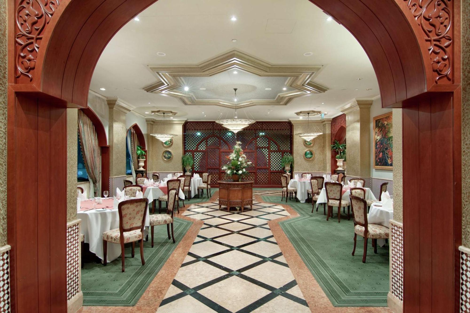 Madinah Hilton Hotel Medina Exterior photo The photo shows an elegantly designed restaurant interior. There is an arched entrance that leads into the dining area. The space features several round tables, each set with white tablecloths and fine diningware. The flooring has a geometric pattern