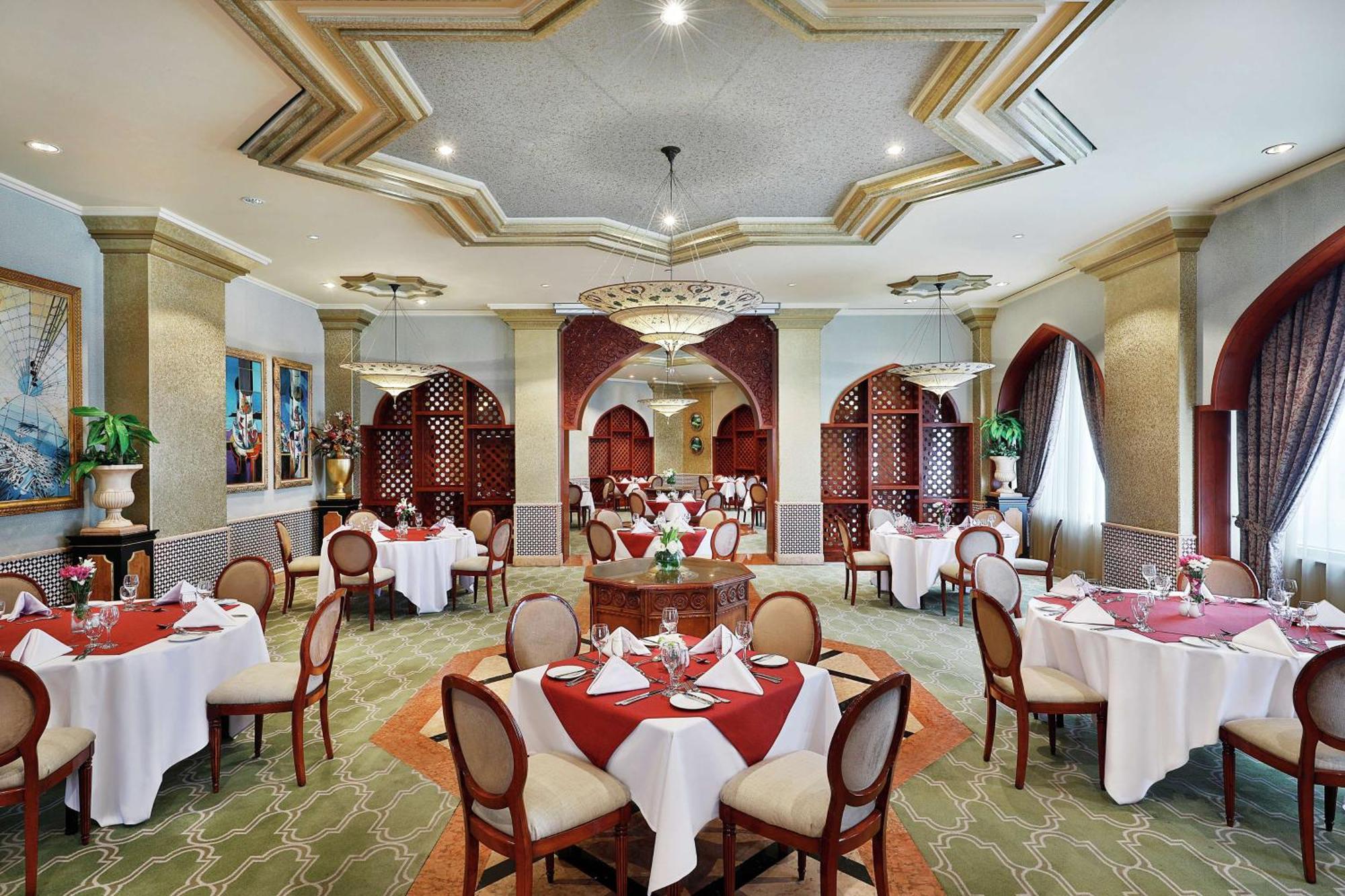 Madinah Hilton Hotel Medina Exterior photo The photo shows an elegant restaurant interior. It features well-arranged dining tables, each set with white tablecloths, red napkins, and tableware, creating a refined atmosphere. The room has decorative elements like intricate ceiling patterns and 