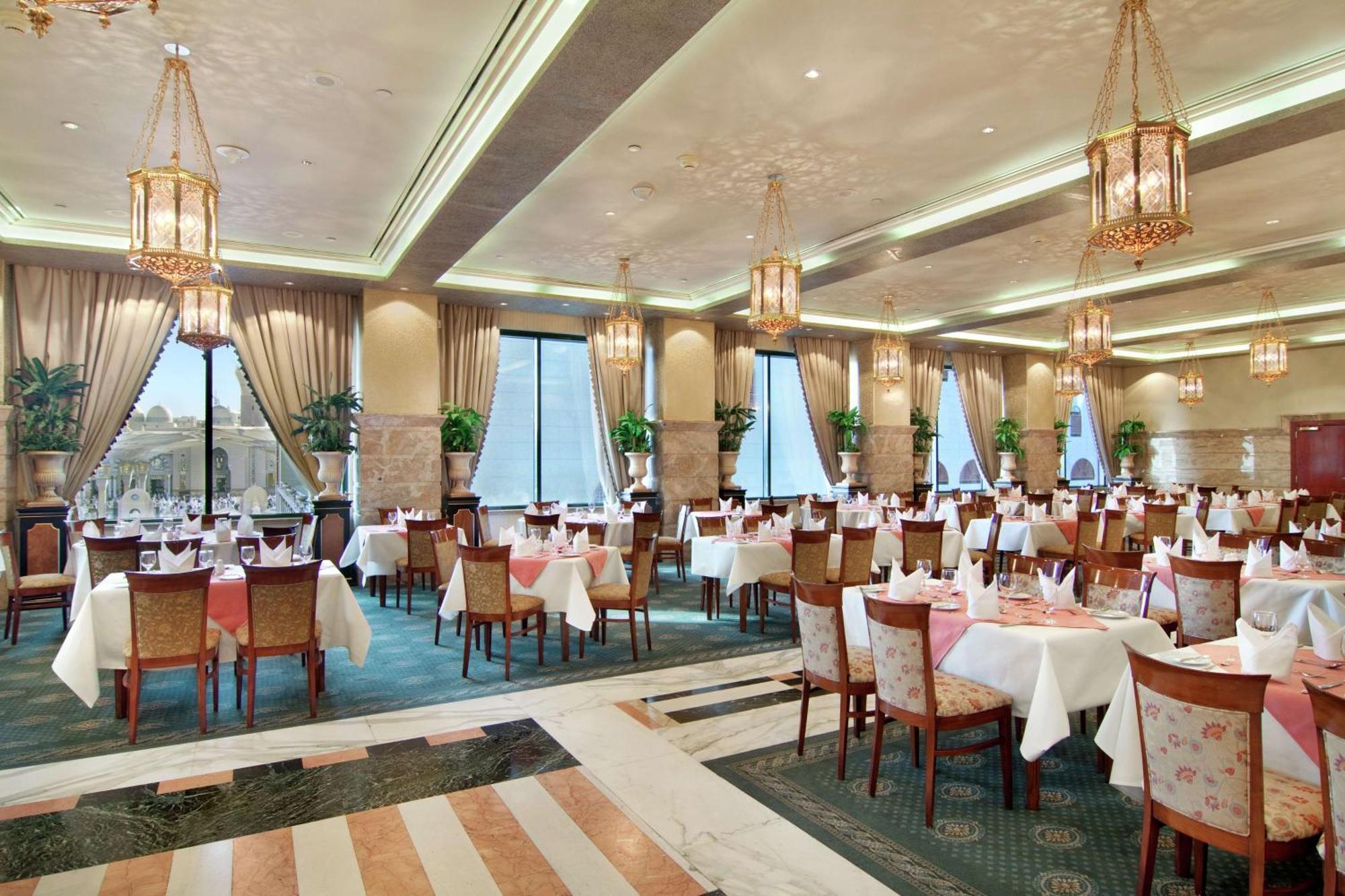 Madinah Hilton Hotel Medina Exterior photo The photo shows an elegant restaurant interior. It features a spacious dining area with tables set for guests, each covered with white tablecloths. Beautifully arranged chairs complement the tables. The decor includes large windows allowing natural l