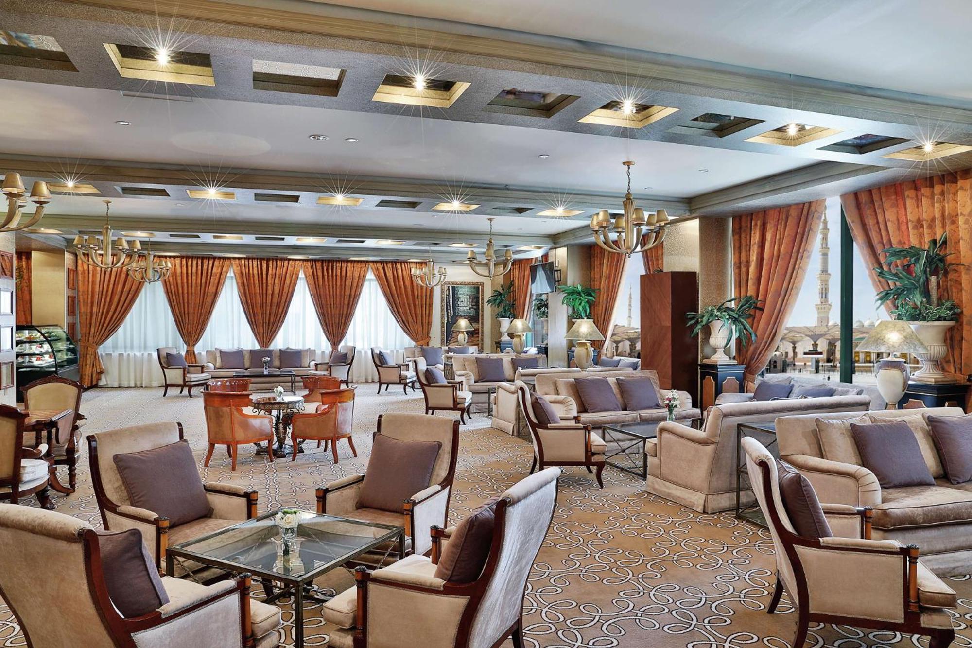 Madinah Hilton Hotel Medina Exterior photo The photo shows an elegant lounge or lobby area. There are several comfortable armchairs and sofas arranged around small tables. The decor features rich curtains in shades of orange and brown, complemented by light-colored walls and a patterned carpe