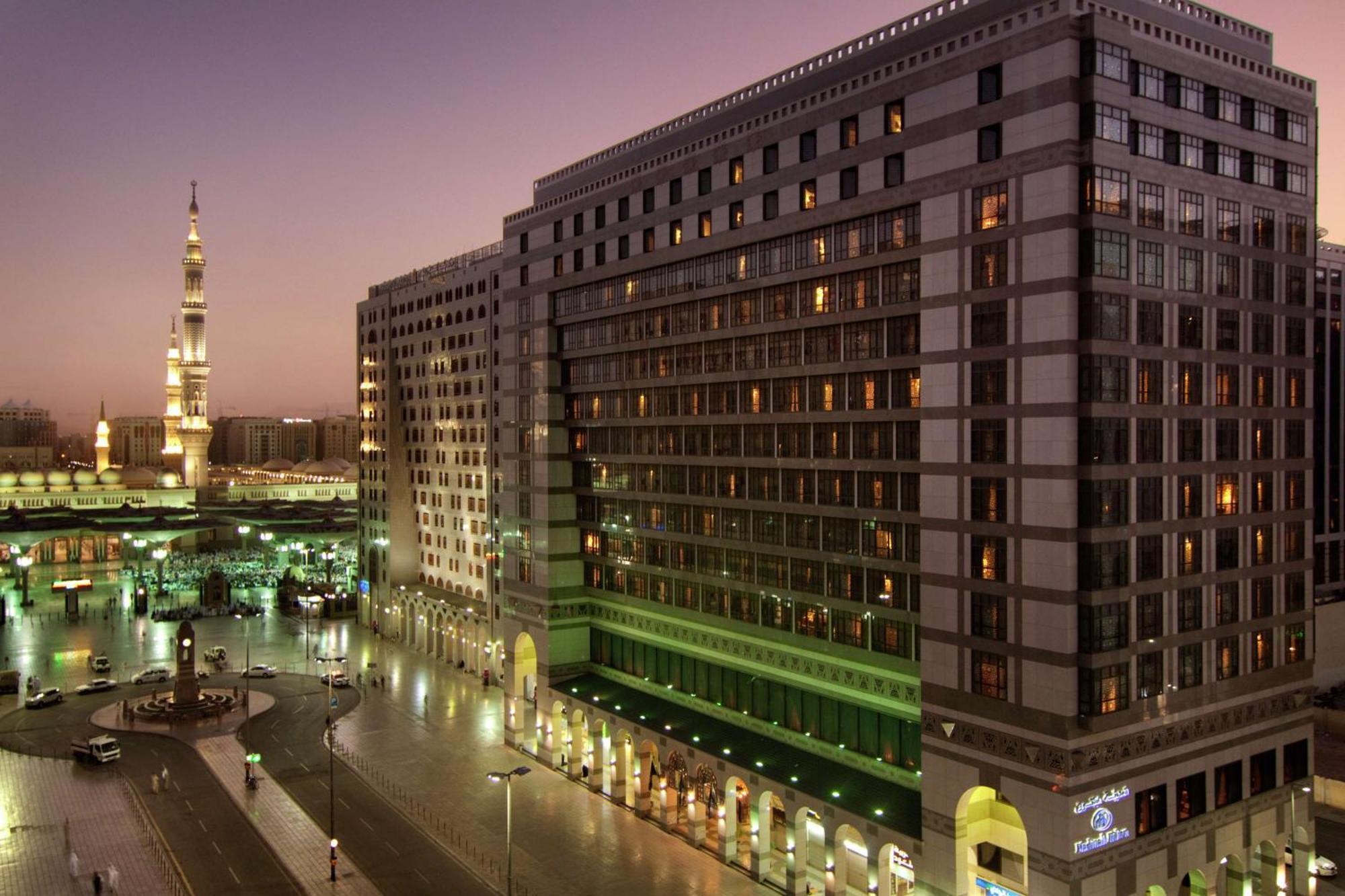 Madinah Hilton Hotel Medina Exterior photo The photo shows a scene of a modern building, likely a hotel, located in a bustling area. The structure features multiple floors with large windows, and its exterior is well-lit. In the background, there is a mosque with a tall minaret, indicating th