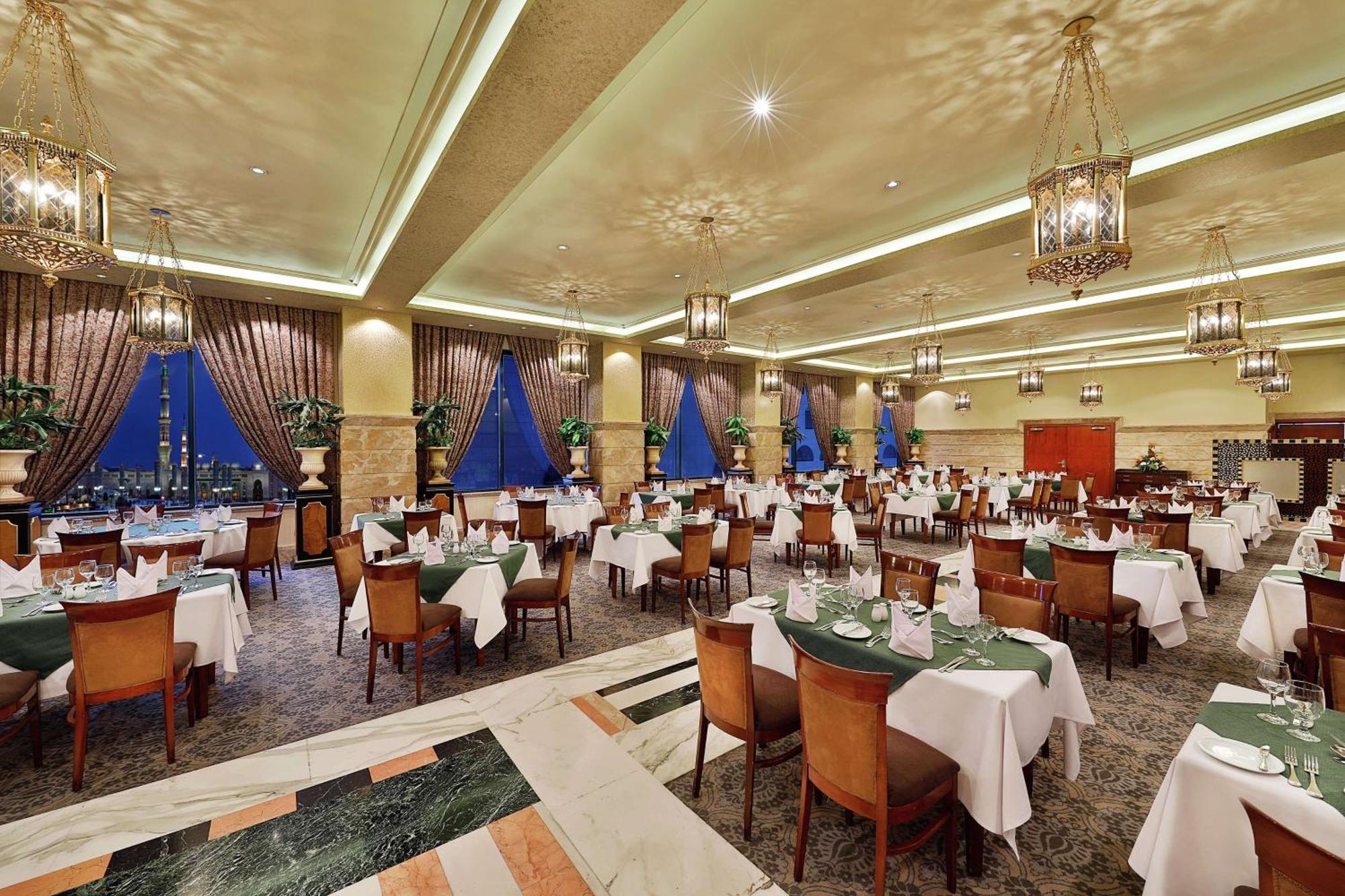 Madinah Hilton Hotel Medina Exterior photo The photo depicts a spacious, elegantly designed dining area of a restaurant. It features numerous tables, each set with white tablecloths and green napkins, along with chairs arranged neatly. The decor includes large windows draped with curtains, al